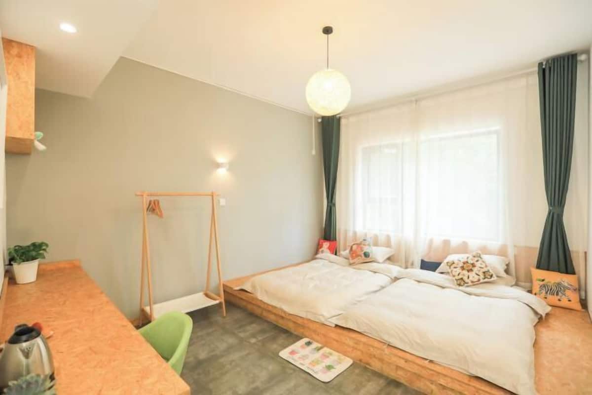 A bedroom with a wooden bed, a desk, a chair, a hanging light fixture, and a window with curtains.