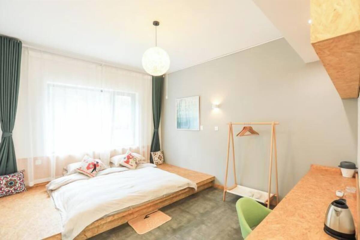 A bedroom with a bed, a desk, a chair, a hanging coat rack, and a window with curtains.