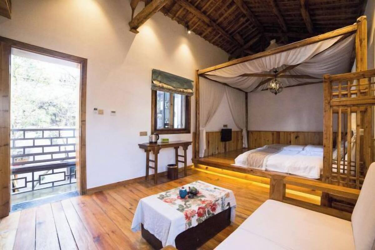 A room with a wooden ceiling, a bed with a canopy, a small table with a floral tablecloth, and a window with a view of greenery.