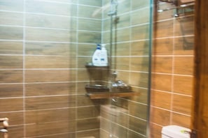 Et badeværelse med fliserede vægge, en glasbrusekabine og en hylde med toiletartikler.