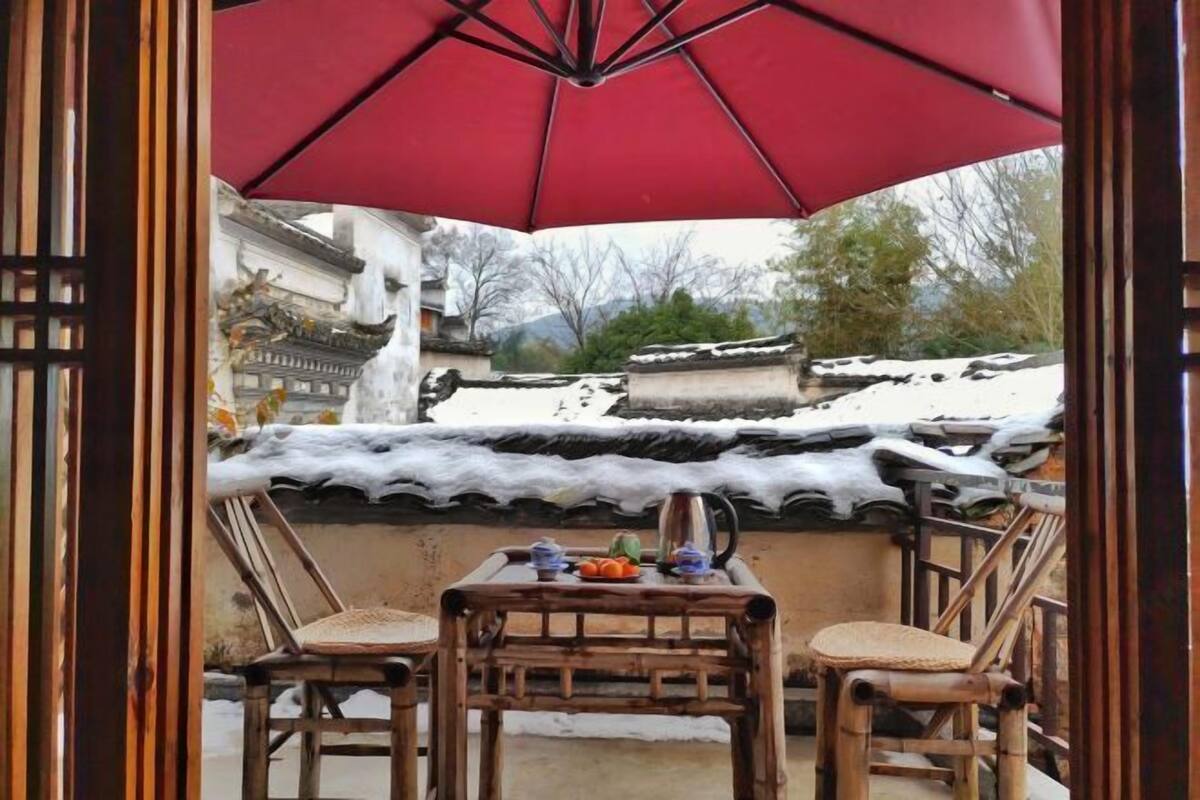 A traditional tea set on a table under a red umbrella, with a view of a snow-covered building in the background.