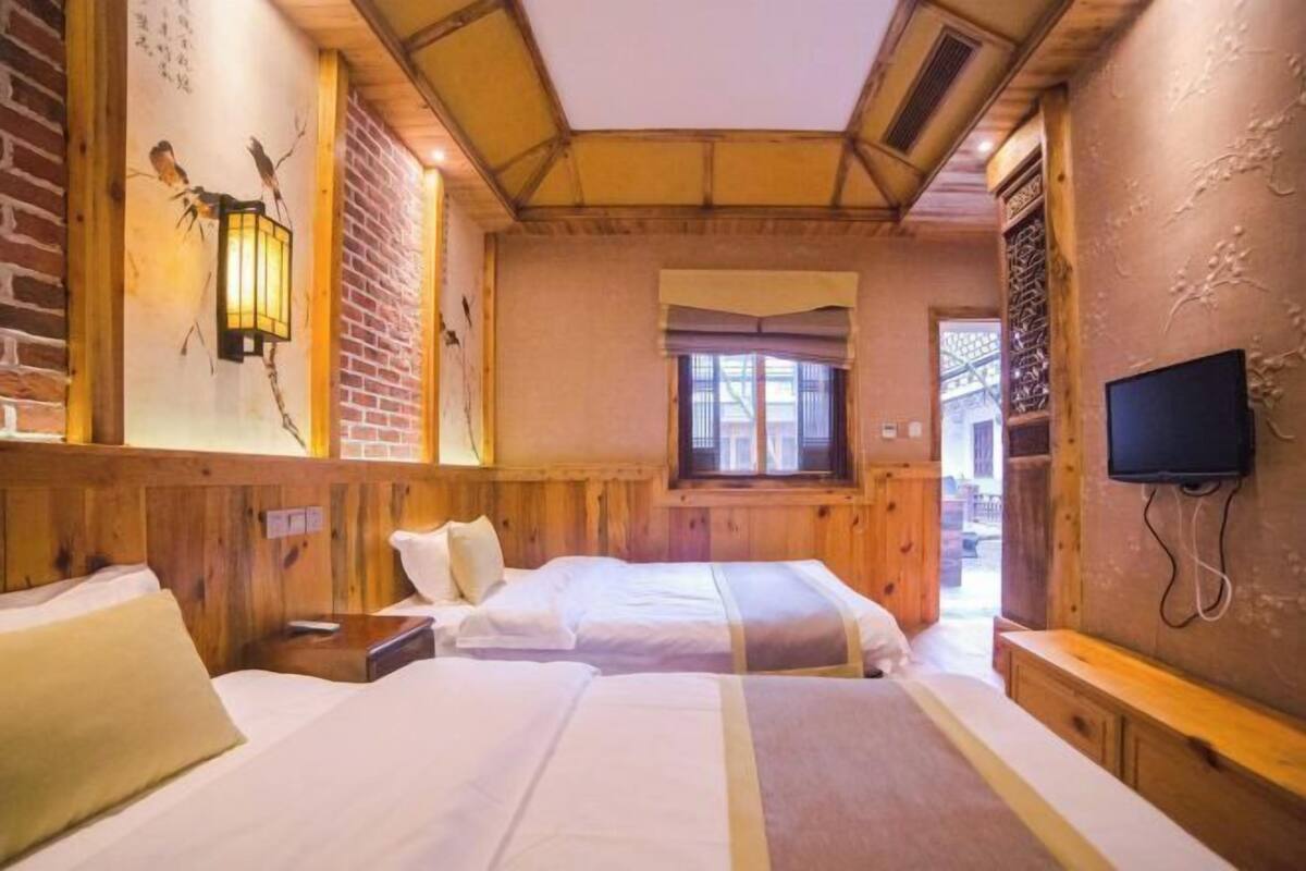 A room with wooden walls, a bed, a bedside table, a lamp, a window, and a TV.