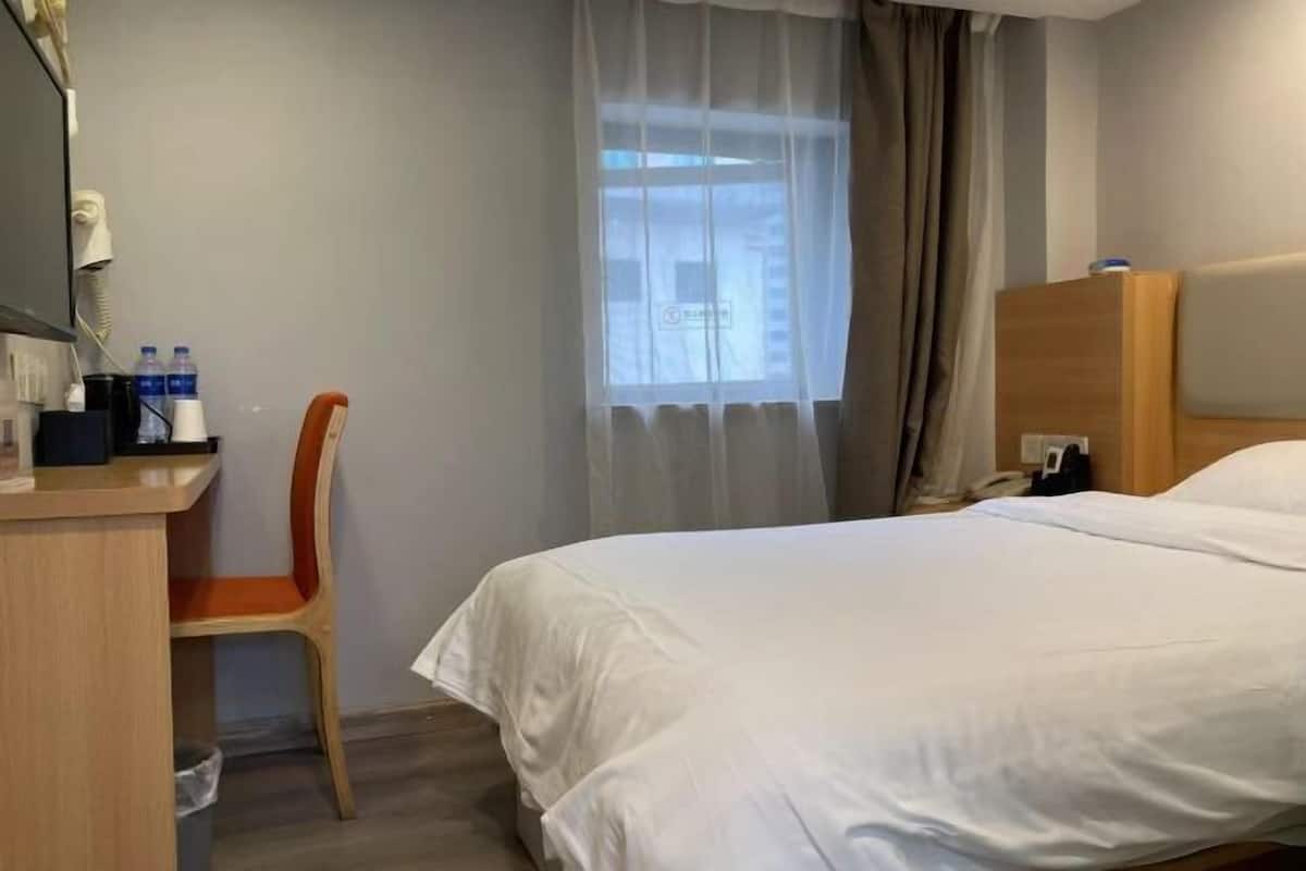 A hotel room with a bed, a desk, a chair, a window with curtains, and a wooden headboard.