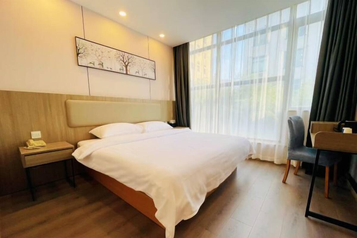 A hotel room with a large bed, a bedside table with a lamp, a chair, and a desk with a chair.