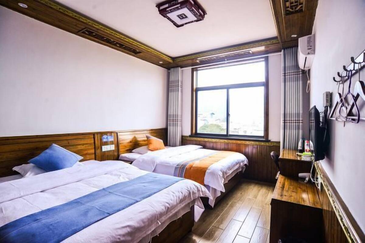 Quarto de hotel com duas camas, cabeceira de madeira, janela com cortinas, escrivaninha de madeira e televisão fixada na parede.