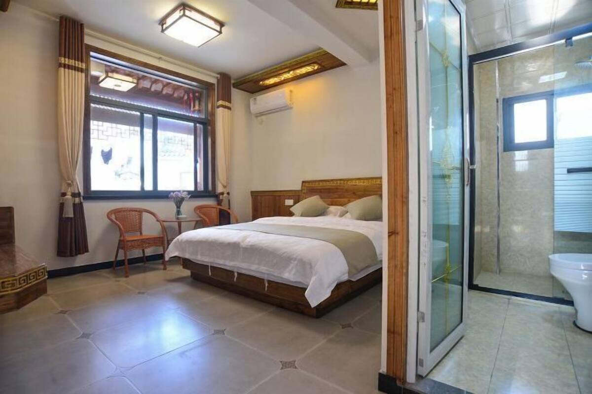 Quarto de hotel com cama, poltrona, mesa, janela com cortinas, ar condicionado e chuveiro em gabinete de vidro.