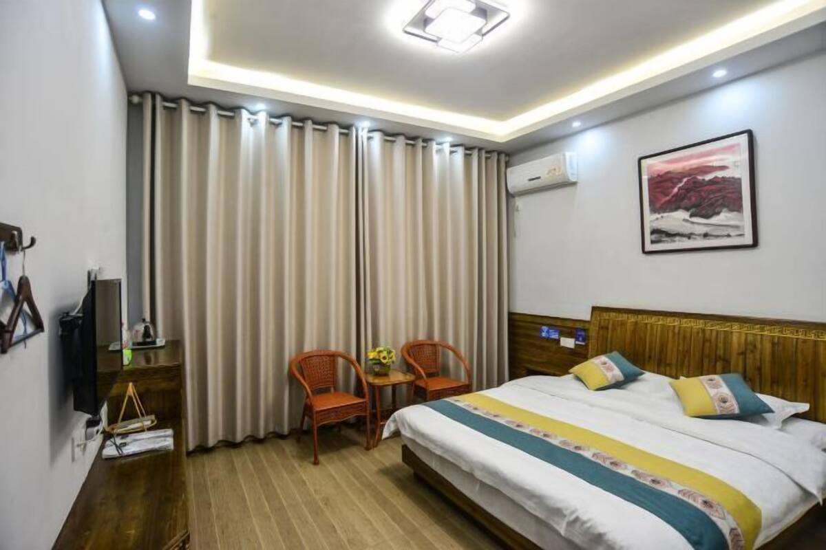 Quarto de hotel com uma cama, duas cadeiras, uma mesinha com um vaso, um quadro na parede e uma luminária no teto.