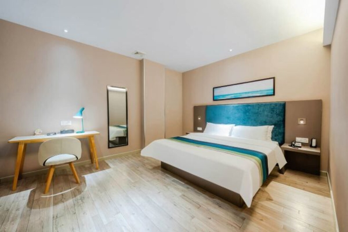 A hotel room with a bed, a desk, a chair, a nightstand, and a mirror.