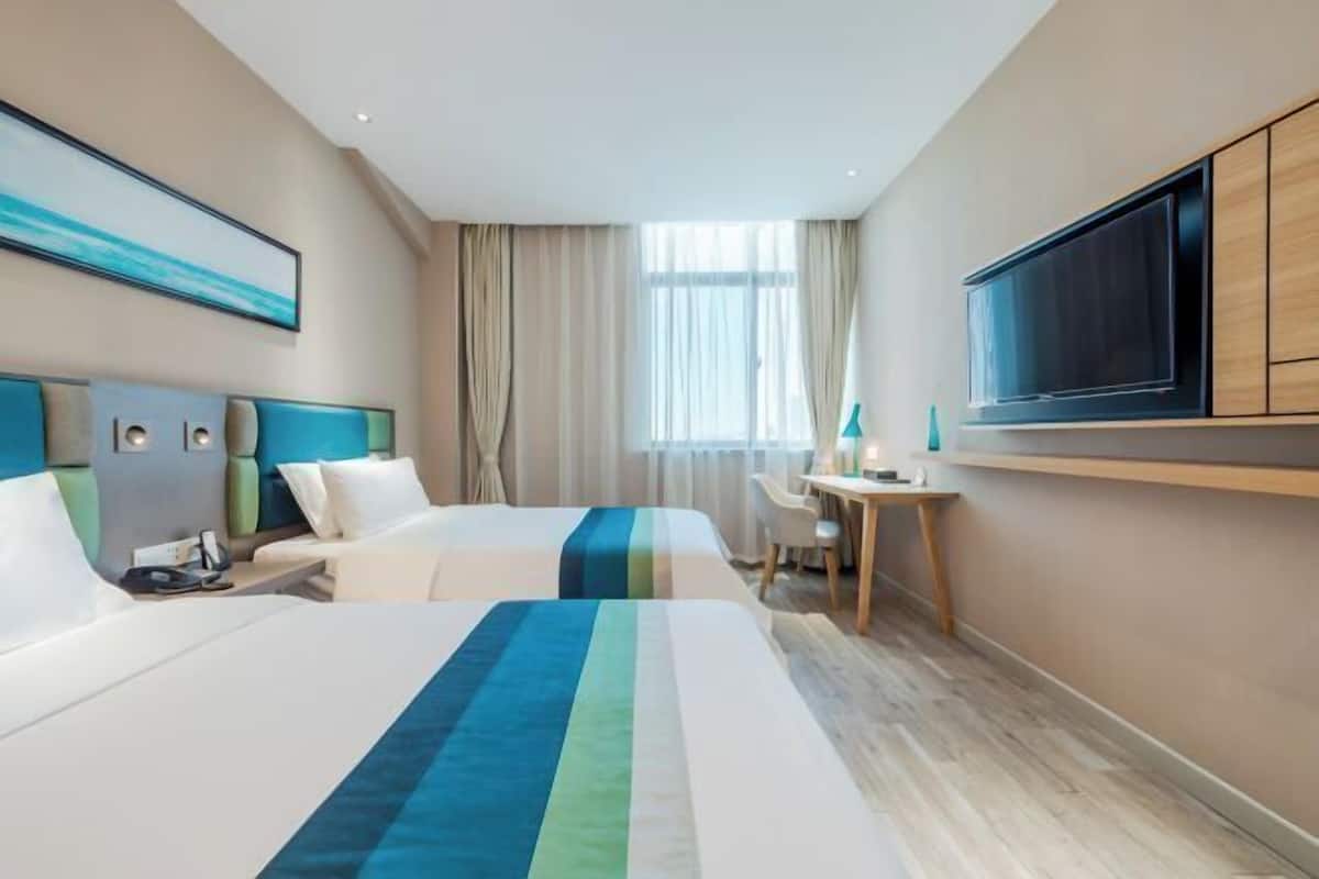 A hotel room with two beds, a desk, a television, and a large window with curtains.