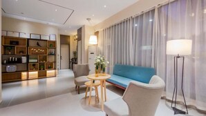 Ein modernes Wohnzimmer mit einem blauen Sofa, zwei Sesseln, einem kleinen runden Tisch und einer Stehlampe. Ein Bücherregal beherbergt verschiedene Gegenstände, und ein großes Fenster ist mit durchsichtigen Vorhängen drapiert.