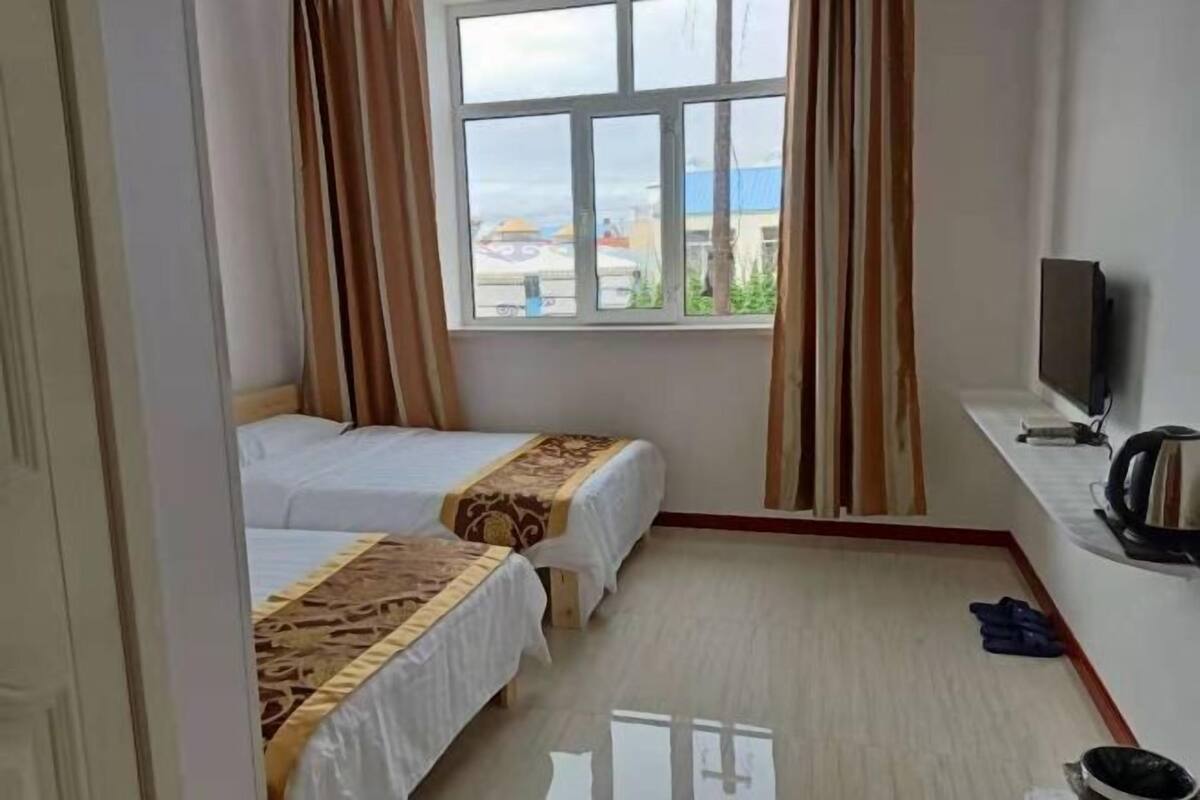 Ein Zimmer mit zwei Betten, einem Fenster mit Vorhängen, einem Fernseher und einem kleinen Tisch mit Wasserkocher.