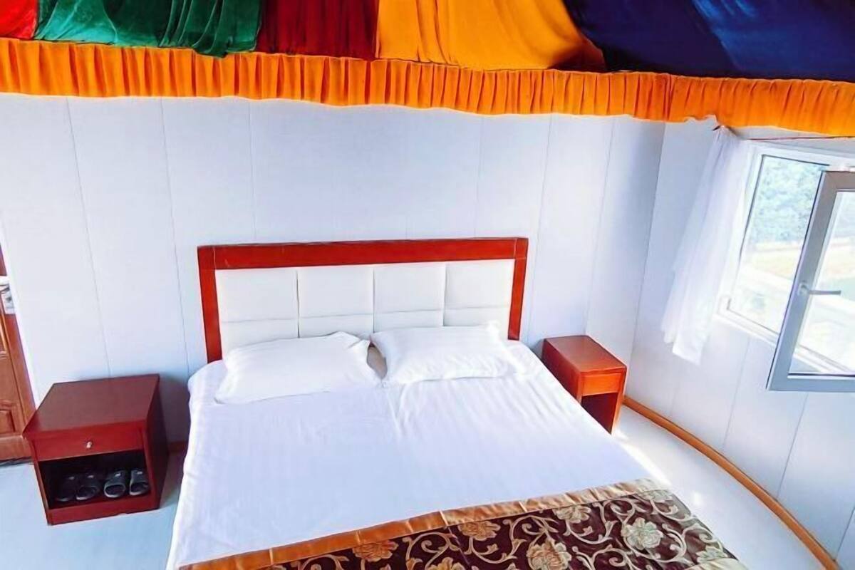 Ein ordentlich bezogenes Bett mit weißen Laken und einer braunen, gemusterten Tagesdecke, an dessen Seiten Holz-Nachttische mit Schubladen stehen, in einem Zimmer mit weißen Wänden und einem Fenster mit Vorhängen.