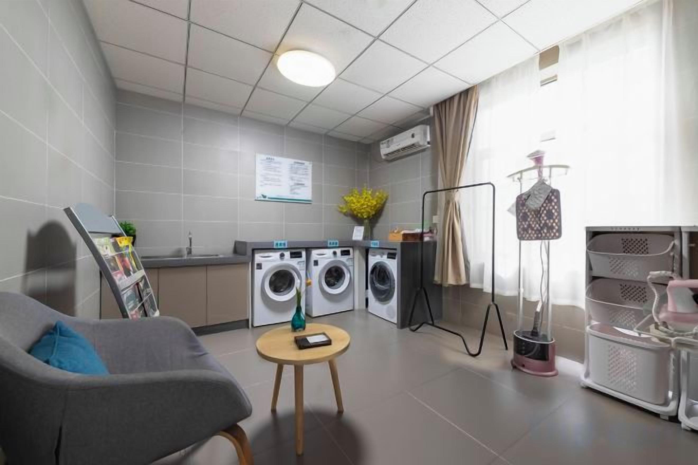 A compact hotel room with a grey sofa, a small round table, a washing machine, and a drying rack.
