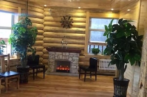 Interior de una cabaña de troncos con chimenea de piedra, suelo de madera y plantas en macetas.