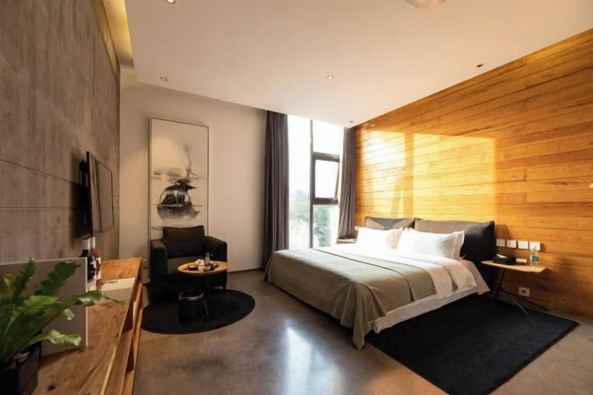 A modern hotel room with a bed, a seating area, a TV, and a large window.