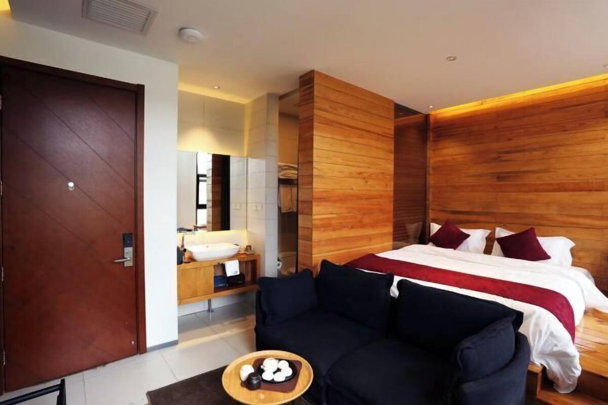 A modern hotel room with a wooden accent wall, a bed with red and white bedding, a black sofa, a wooden side table, and a small round table with a bowl of fruit.