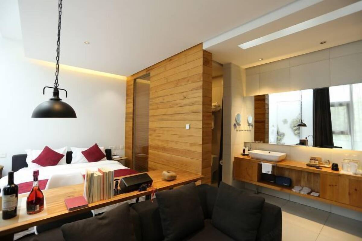 A modern hotel room with a wooden accent wall, a large bed with red pillows, a desk with books and a laptop, a sofa, and a bathroom with a large mirror and sink.