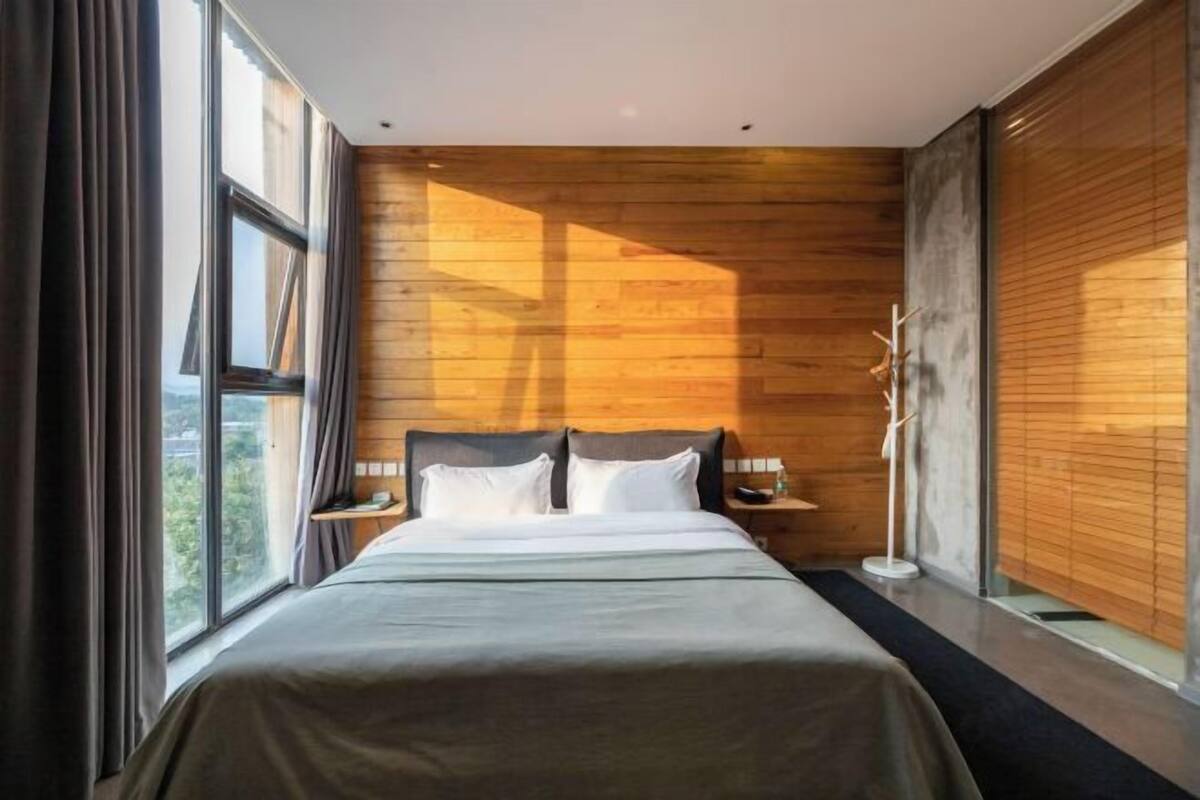 A modern bedroom with a large bed, wooden panel walls, a nightstand, a floor lamp, and a window with curtains.