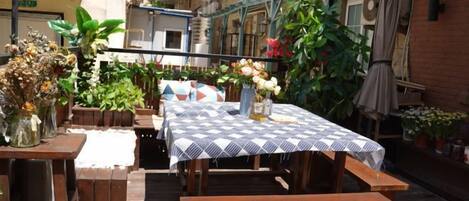 Un patio exterior con una mesa y bancos, rodeado de plantas en macetas y un muro de ladrillo.