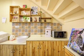 Una cocina con armarios de madera, un lavamanos blanco y un microondas. Se observan estantes con diversos objetos y un cuadro en la pared.
