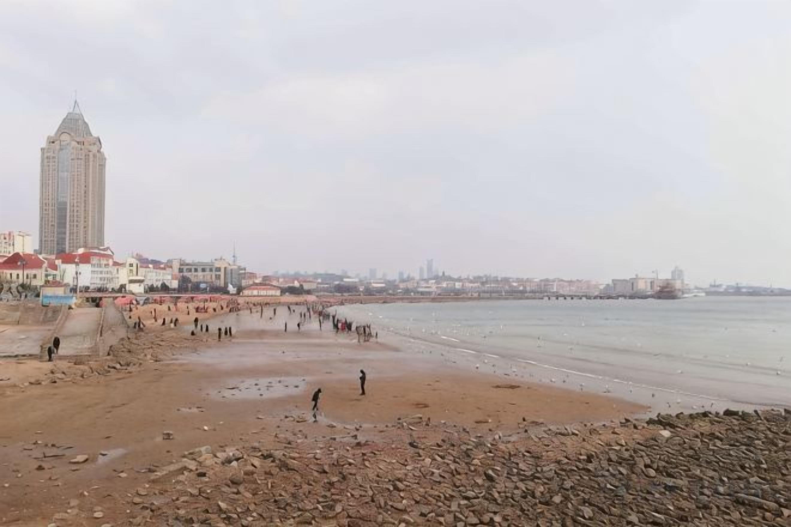 Une plage avec un rivage rocheux, une jetée qui s’avance dans l’eau et des bâtiments en arrière-plan.