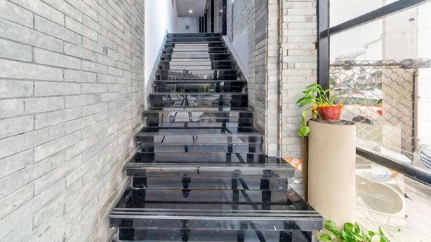 Un escalier aux marches noires menant à un étage supérieur, flanqué de murs en briques gris clair, et un pot de fleurs placé sur la droite.