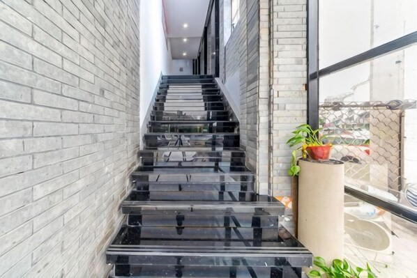Una escalera con escalones negros que ascienden a un piso superior, flanqueada por paredes de ladrillo gris claro, y una planta en maceta a la derecha.