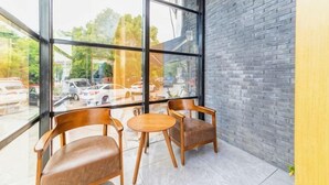 Un coin avec deux chaises en bois et une petite table, de grandes fenêtres donnant sur des voitures garées et des arbres, et un mur en briques.