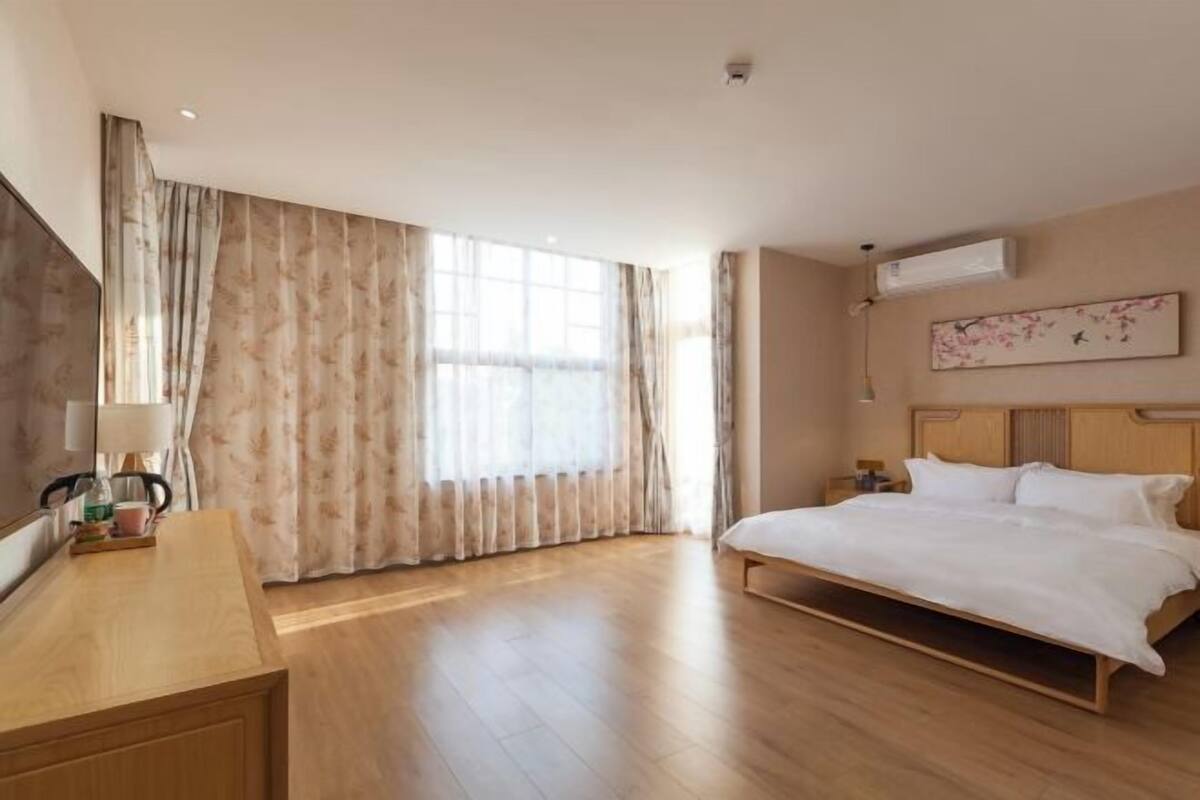 A hotel room with a wooden bed, a desk with a lamp, a large window with curtains, and an air conditioning unit on the wall.