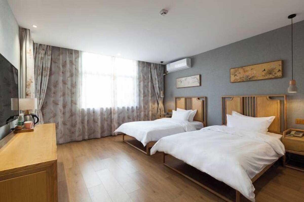A hotel room with two beds, a wooden desk, a window with curtains, a painting on the wall, and an air conditioning unit.