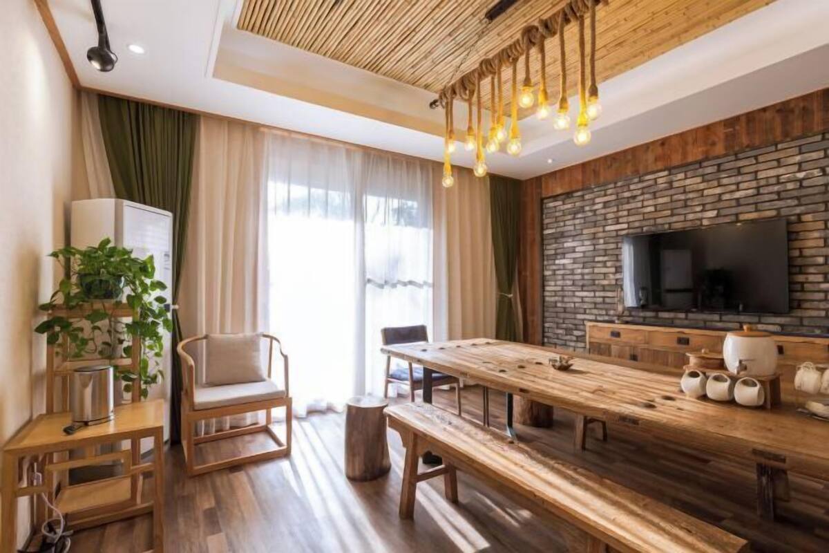 A modern living room with a wooden dining table, a sofa, a TV mounted on a brick wall, and a hanging light fixture.
