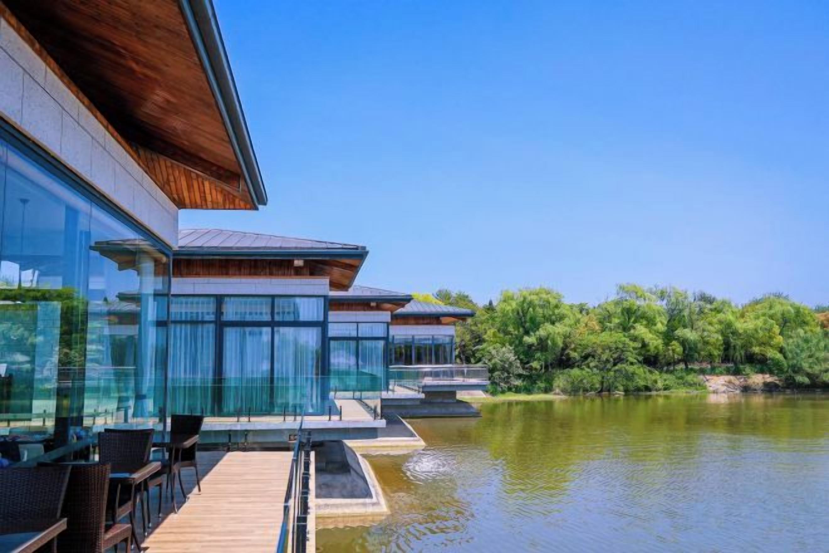 A modern building with a wooden deck overlooking a calm lake, surrounded by lush greenery.