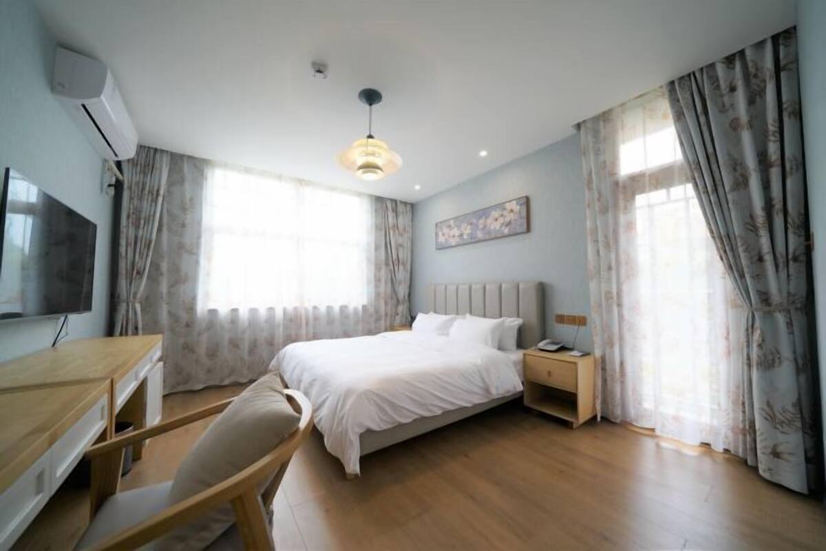 A bedroom with a bed, bedside table, chair, desk, TV, and curtains.
