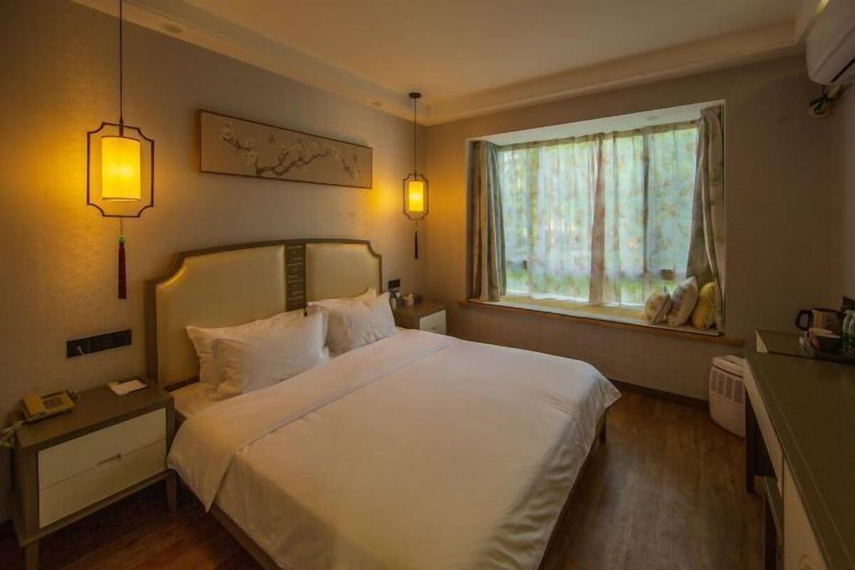 A hotel room with a bed, bedside tables, a window with curtains, a wall-mounted artwork, and a hanging light fixture.