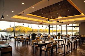 A spacious dining area with multiple tables and chairs, large windows offering a view of a body of water and trees, and pendant lighting.