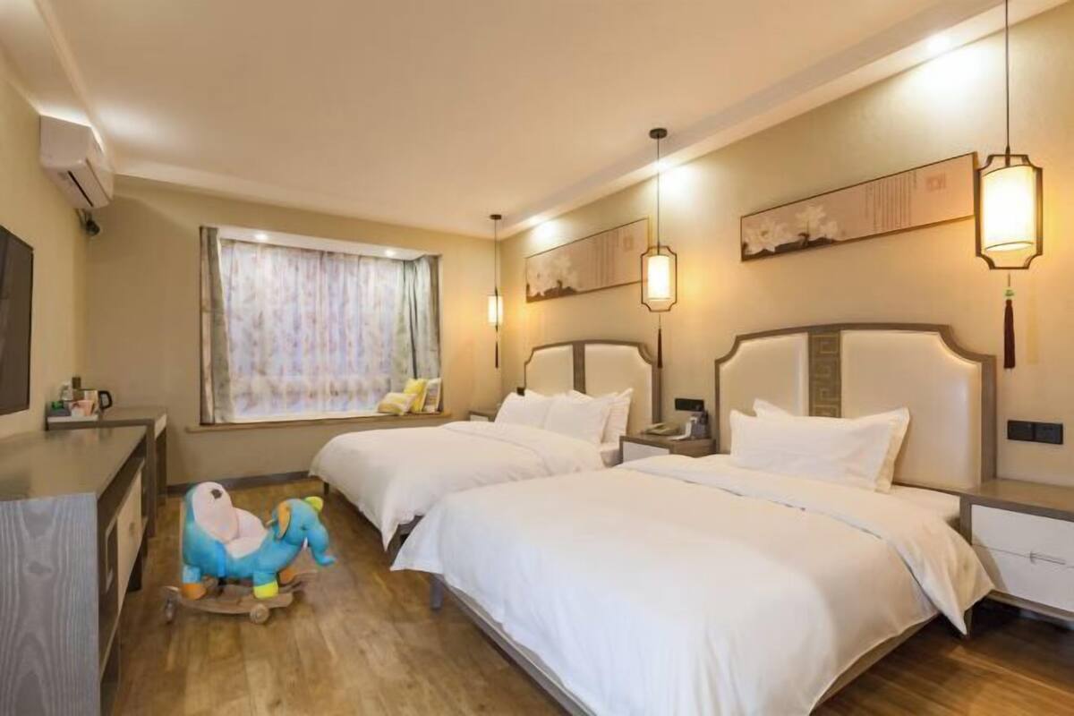 A hotel room with two beds, a desk, a chair, a window with curtains, and a wall-mounted artwork.