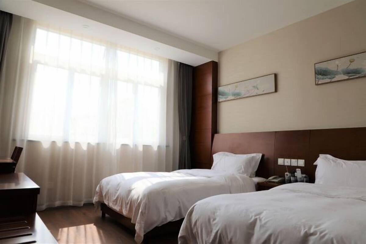 A hotel room with two beds, a desk, a chair, and a window with sheer curtains.