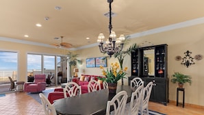A spacious dining area with a dark wooden table and white chairs, a chandelier, and a view of the ocean through large windows.