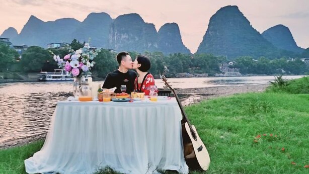 Una pareja besándose en una mesa, con una guitarra, con vistas a un río y montañas.