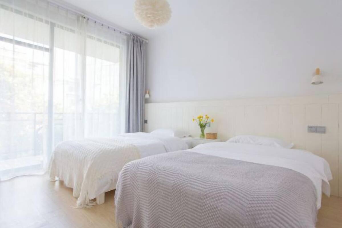 A bright room with two beds, a large window with curtains, and a ceiling light.