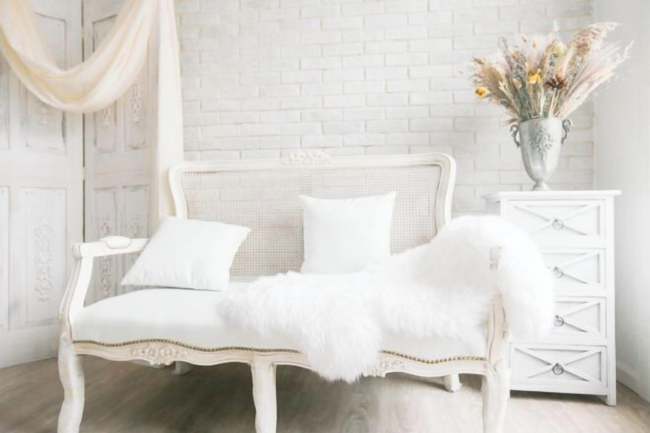 A white vintage sofa with a fluffy white throw blanket, a white dresser with drawers, and a vase with dried flowers on a white brick wall.