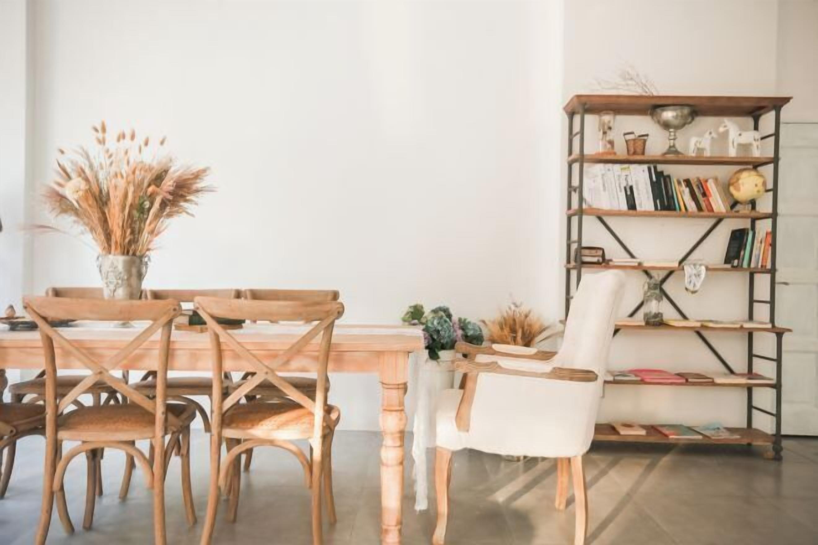 A dining area with a wooden table and chairs, a bookshelf with books and decorative items, and a vase with dried plants.