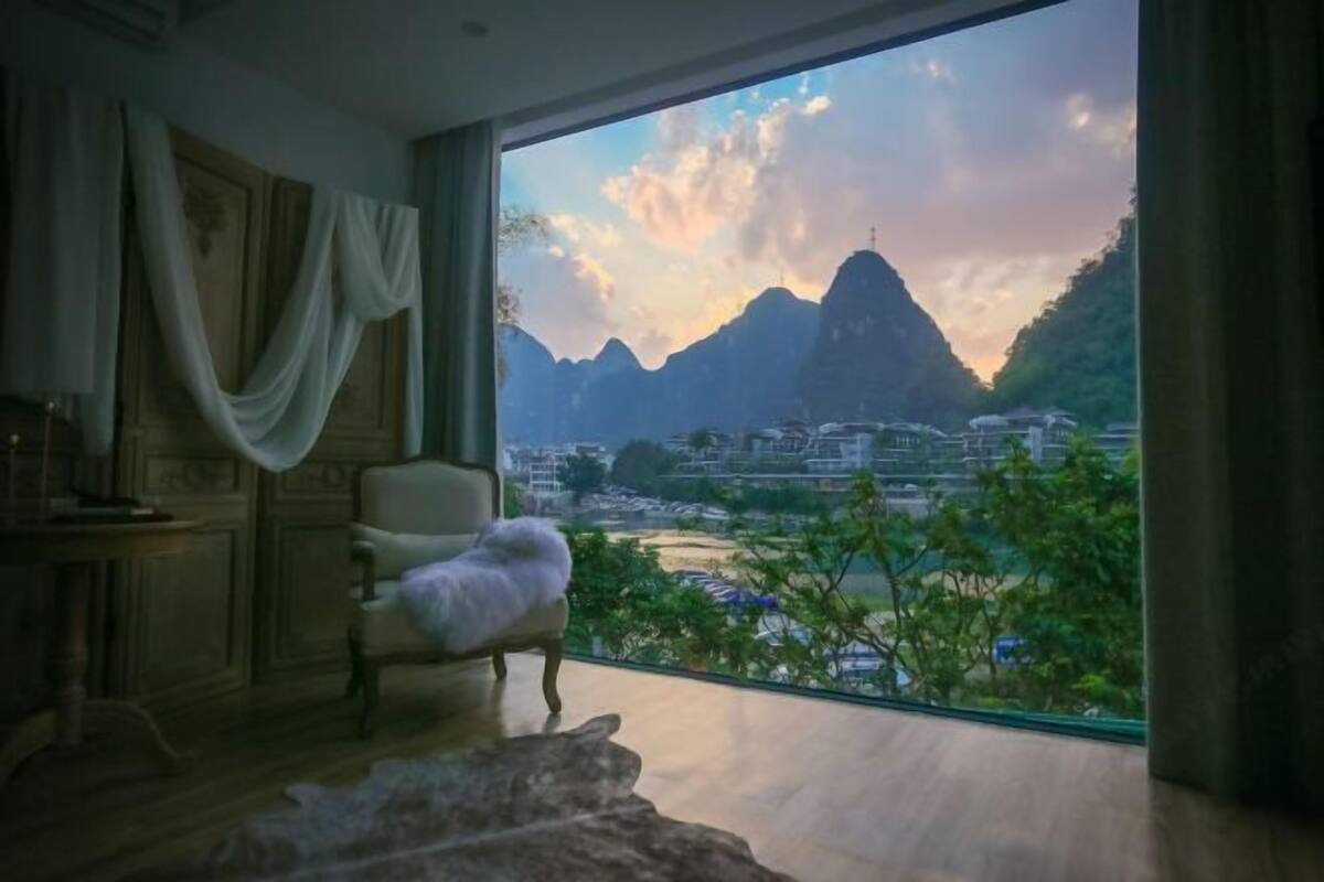 A room with a large window offering a view of mountains and a river. There is a chair with a fluffy cushion and a wooden cabinet.