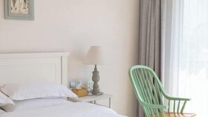 Una cama bien hecha con sábanas blancas, una mesita de noche con una lámpara y una pequeña cesta, y una silla verde cerca de una ventana con cortinas.