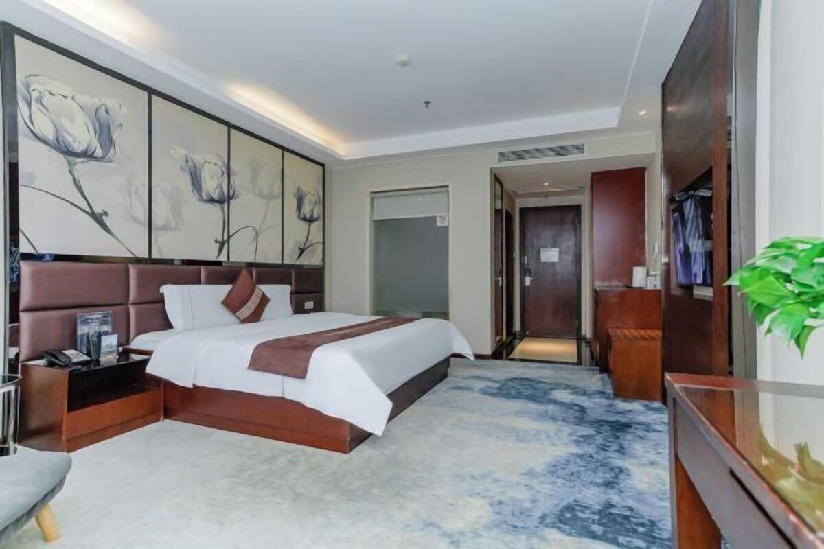 A modern hotel room with a large bed, a nightstand, a bedside table, a wardrobe, and a large window.