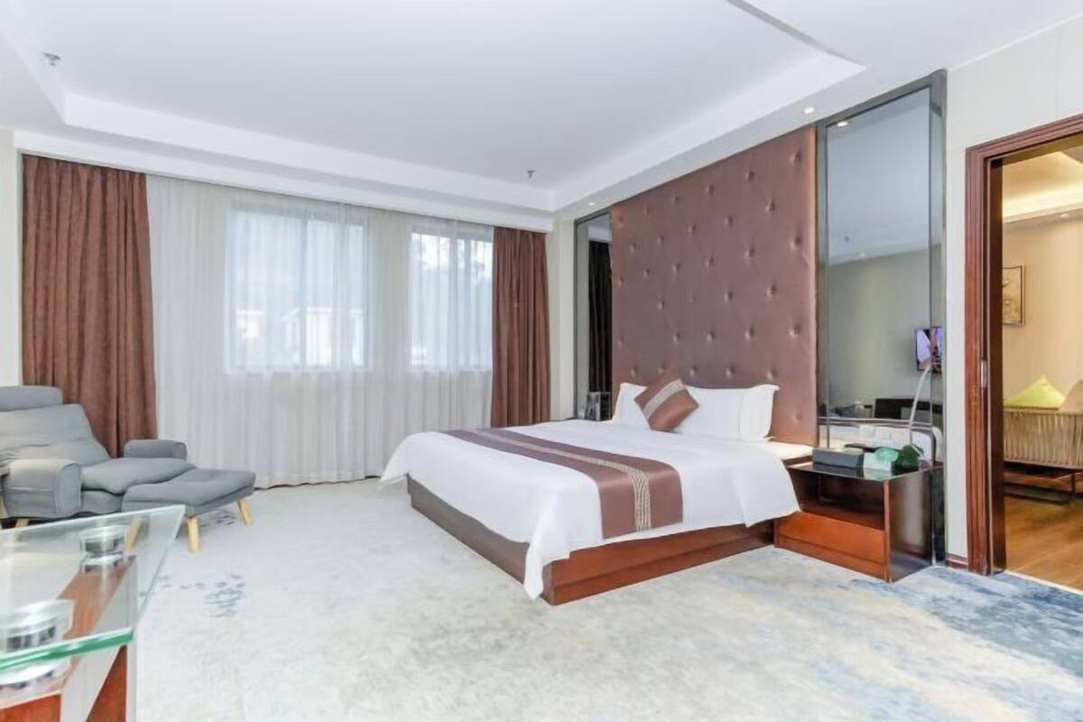 A modern hotel room with a large bed, a seating area with a glass table, and a view through a glass door.