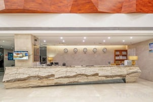 Zona de recepción de hotel con mostrador de mármol, un reloj, mostrador de recepción y una pantalla de televisión.