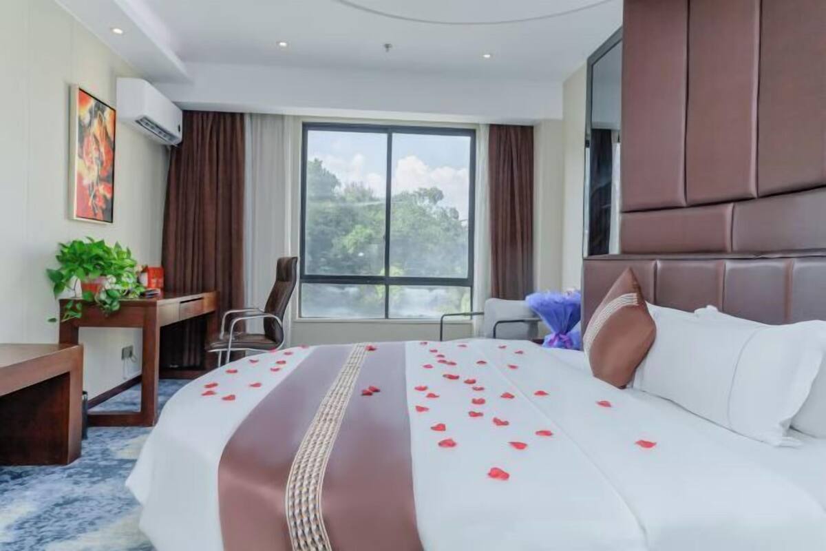 A hotel room with a large bed, a desk, a chair, and a window with a view of trees.