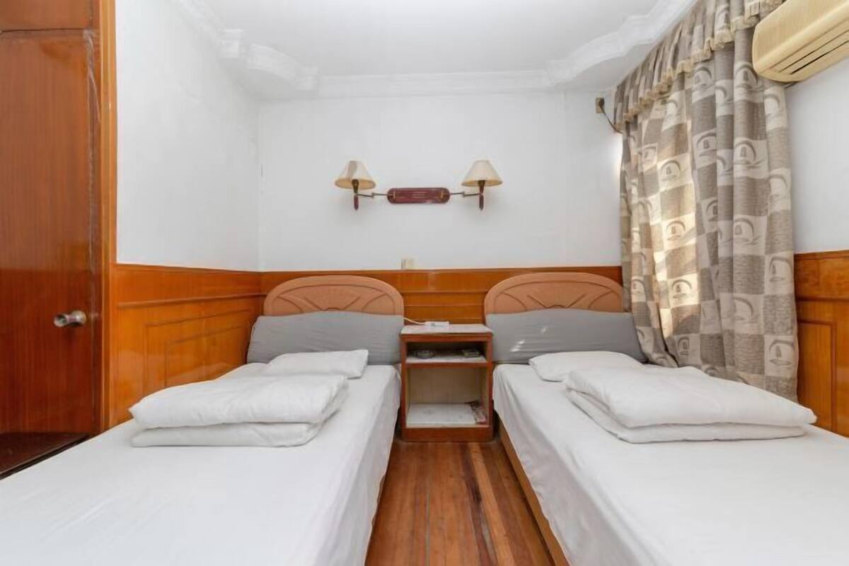 A room with two single beds, a wooden headboard, a nightstand, a wall-mounted lamp, and a patterned curtain.