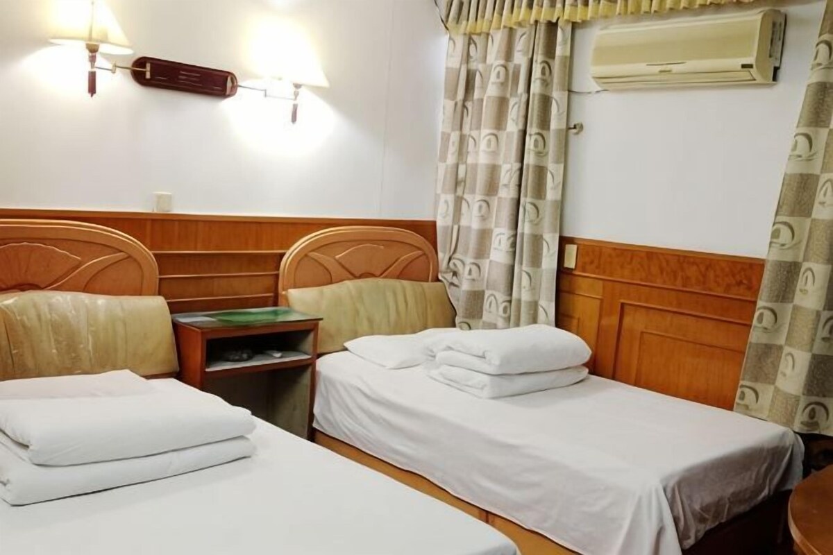 A hotel room with two single beds, a wooden headboard, a nightstand, and a wall-mounted air conditioner.
